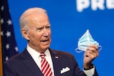 Joe Biden speaks behind a lectern holding a surgical mask.