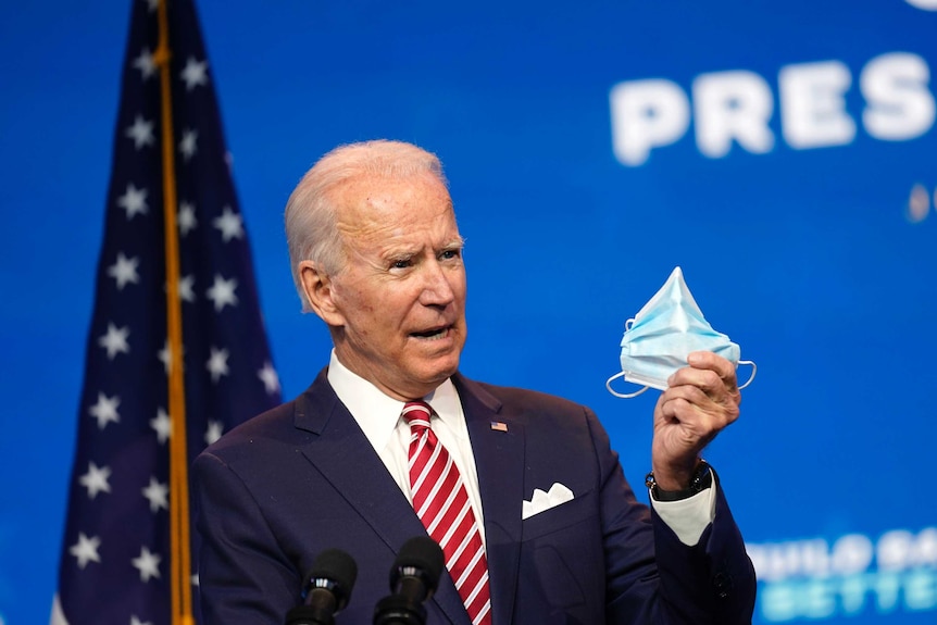 Joe Biden speaks behind a lectern holding a surgical mask.