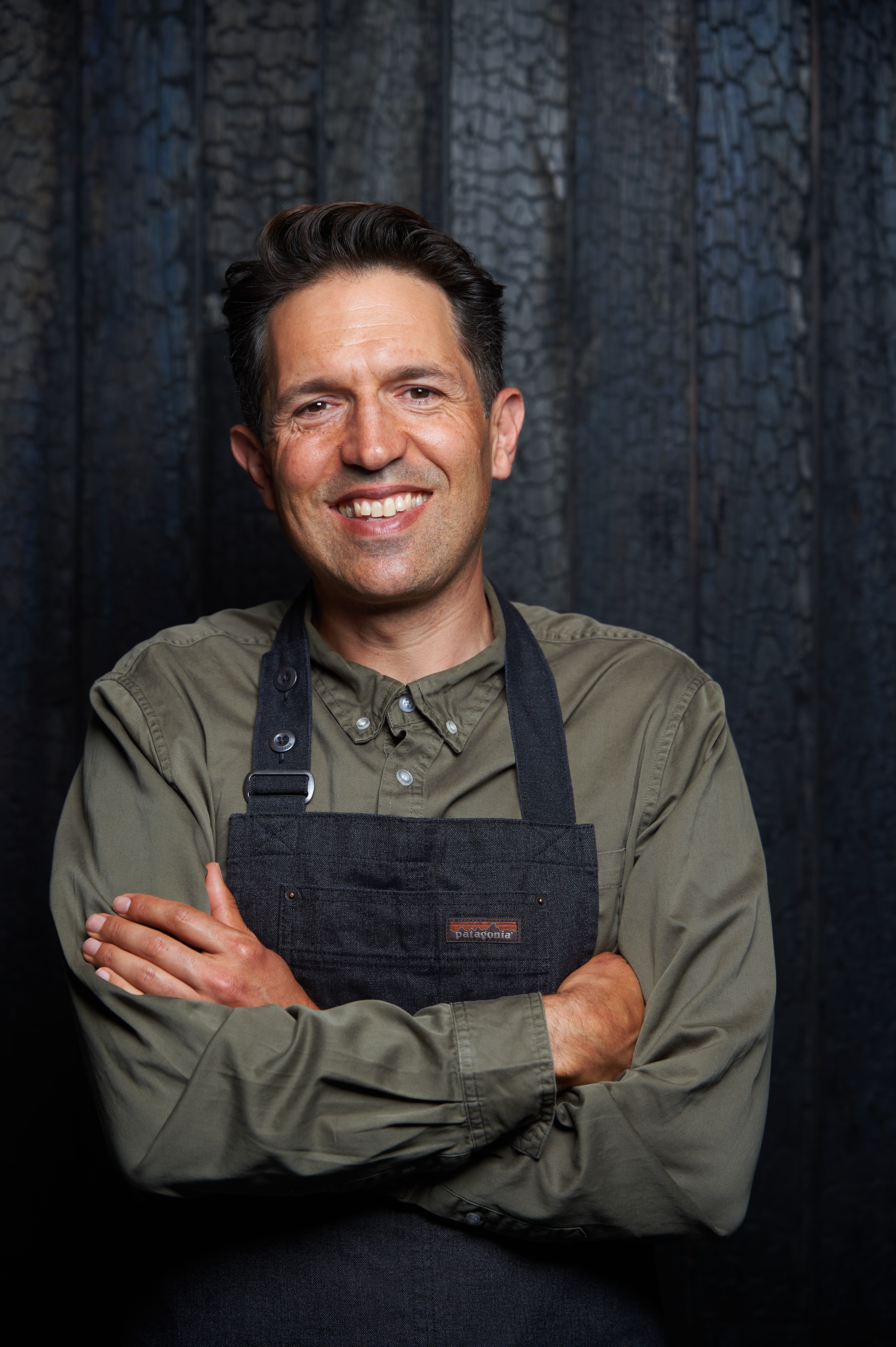 Ben Shewry, smiling widely, with dark short hair and black top, standing against dark wall.
