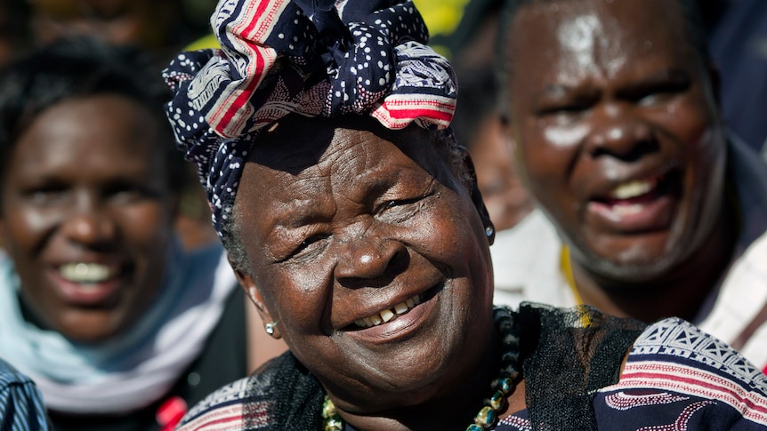 An elderly African woman with a colourful headscarf smiles on a sunny day with African people behind.