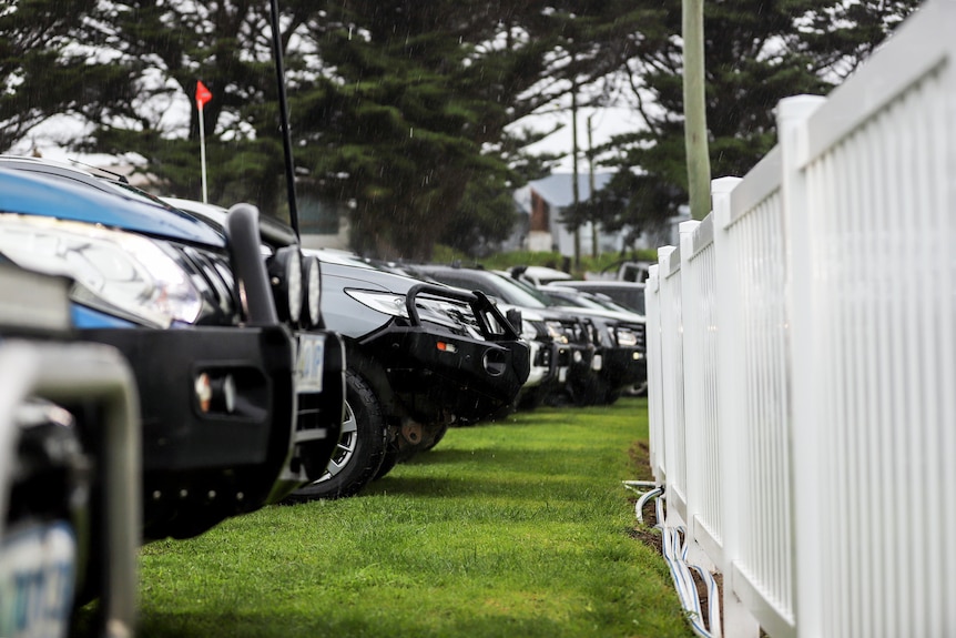 A row of cars, mainly utes, next to a plastic white fence of a football ground.