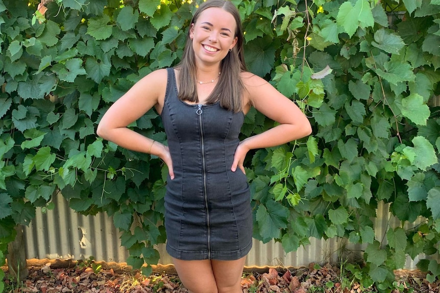 A young woman with brown hair and a black dress smiles in front of fence covered in plants.