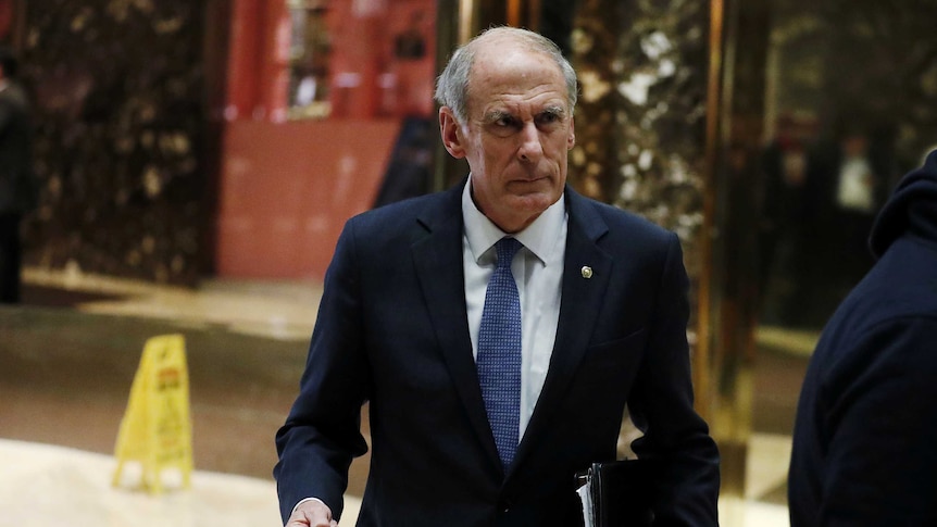 Dan Coats, dressed in a suit, walks with folders and documents in one hand.