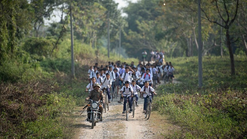 Kids on bicycles follow a man on a motorbike.