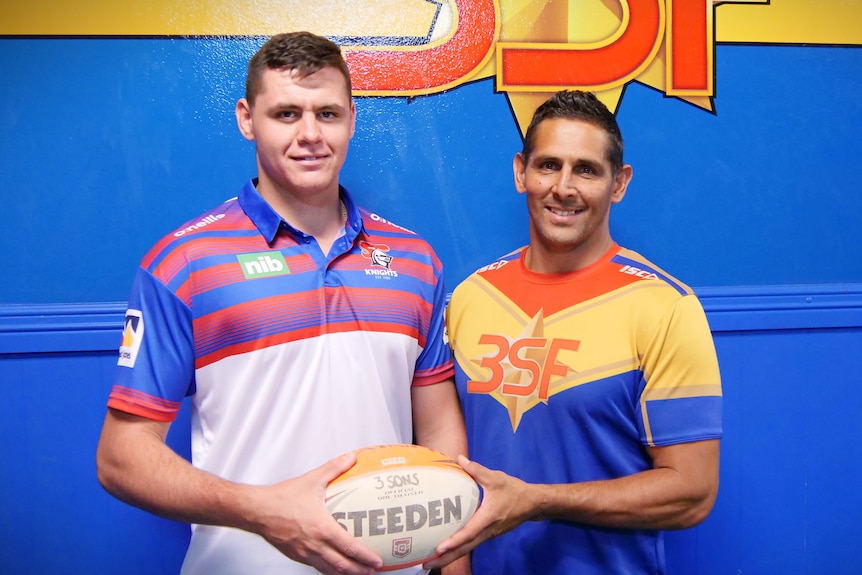two men standing in front of a blue wall holding a rugby ball