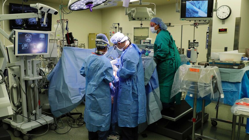 Doctors, wearing blue gowns, operate on a patient in a bed in a hospital ward.