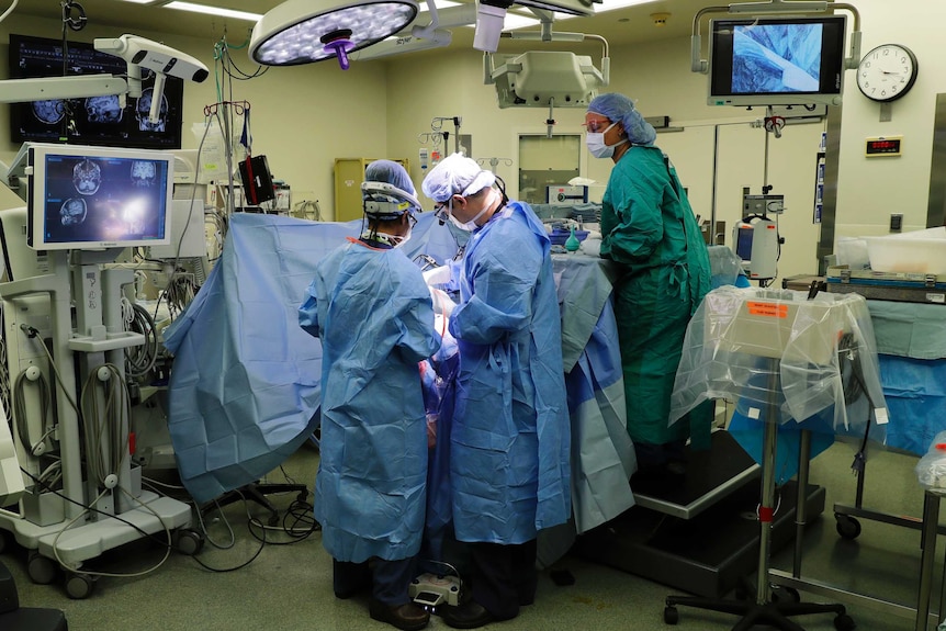 Doctors, wearing blue gowns, operate on a patient in a bed in a hospital ward.