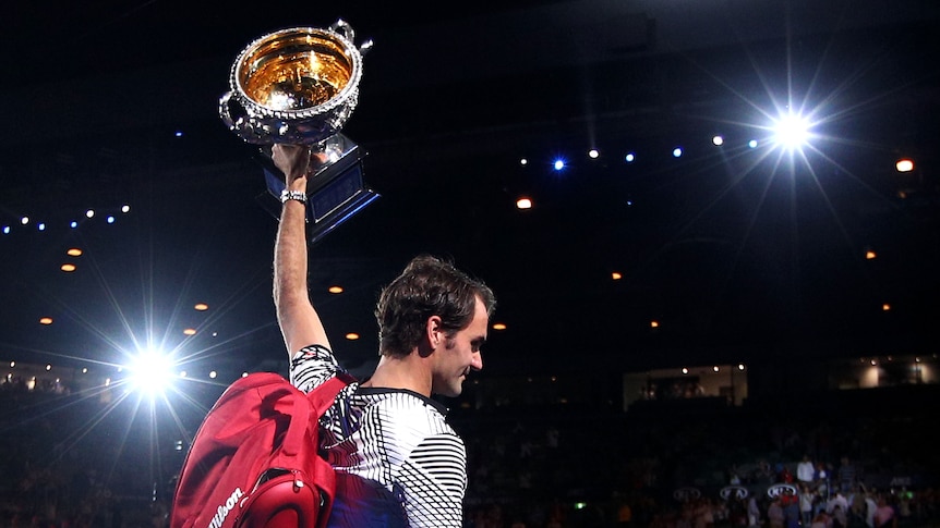 Roger Federer raises the Australian Open trophy in one hand as cameras flash at night while he walks off the court.