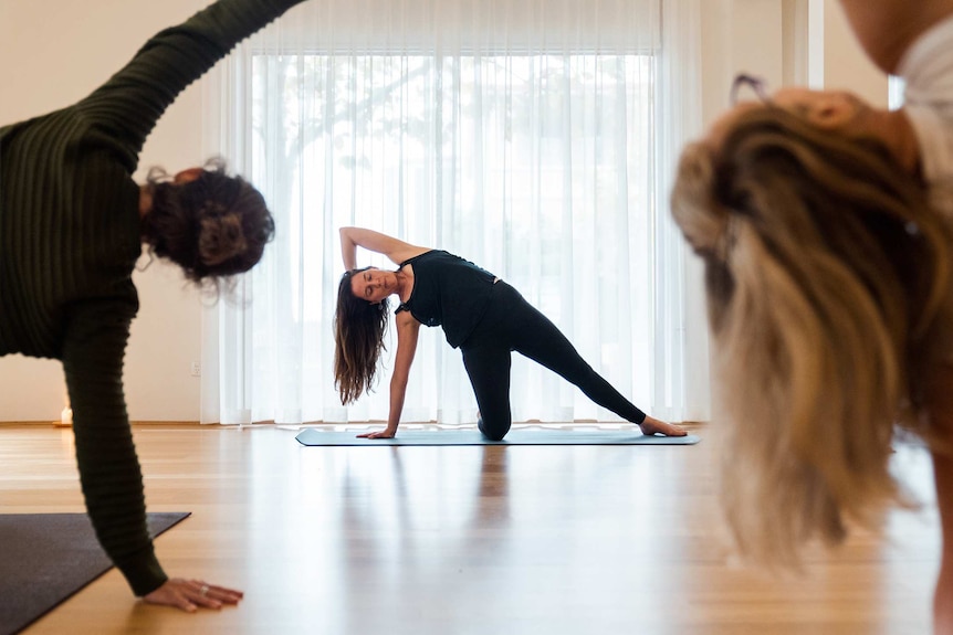 A woman forms a yoga pose at the front of a studio with other class participants following her lead in the foreground.