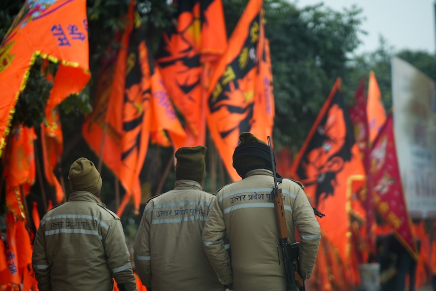 Three people in matching security uniforms, one carrying a weapon, walk past orange flags