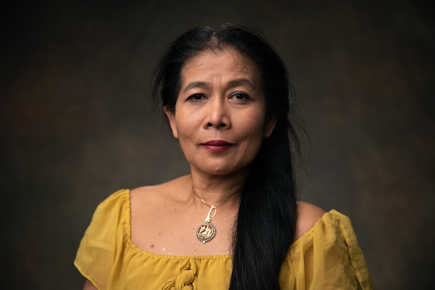 A Thai woman wearing a yellow top with long brown hair looks at the camera. She's in front of a dark brown background