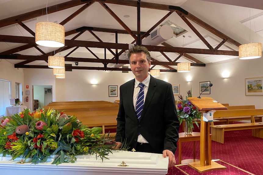 Launceston funeral director Mark Graham standing beside a white coffin with flowers on top of it.