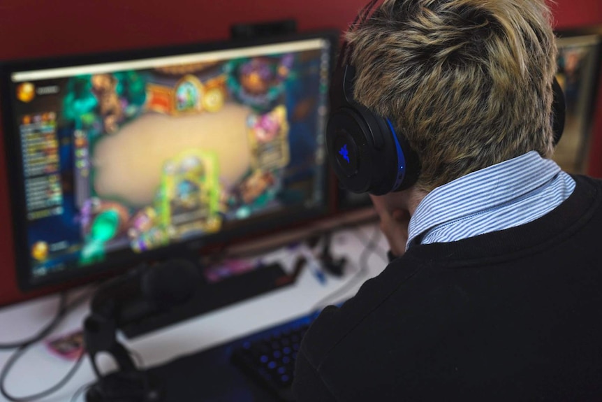 An over-the-shoulder shot of Alex Ridley wearing headphones while playing Hearthstone at a computer.