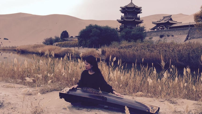 Mindy Meng Wang sits at a guzheng on a riverbank in China. There a stone wall and pavillion in the background.