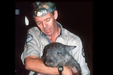 A man wearing a khaki shirt holds a wombat which is asleep