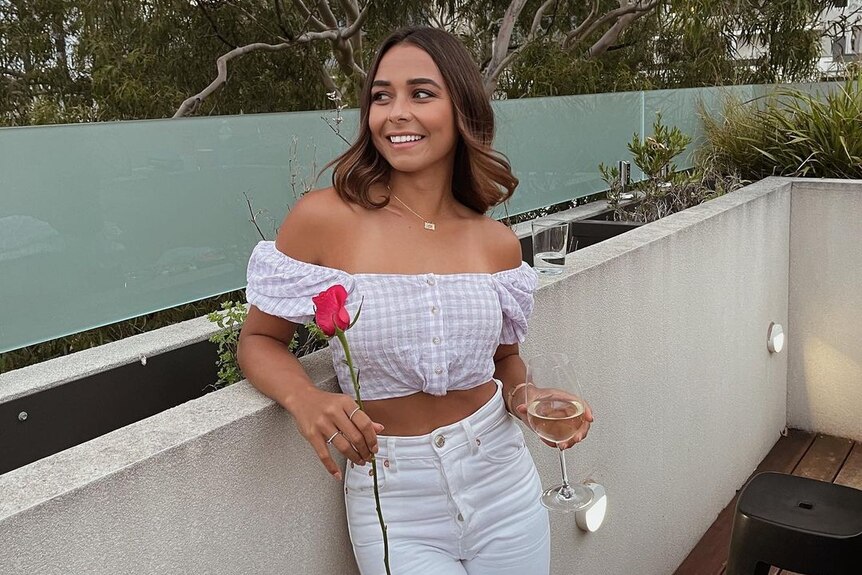 Brooke Blurton poses on a balcony, holding a glass of wine and a rose.