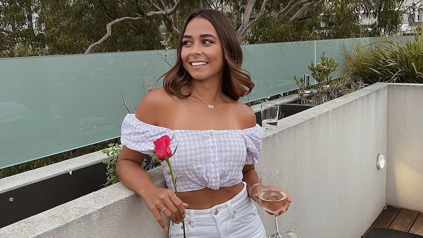 Brooke Blurton poses on a balcony, holding a glass of wine and a rose.