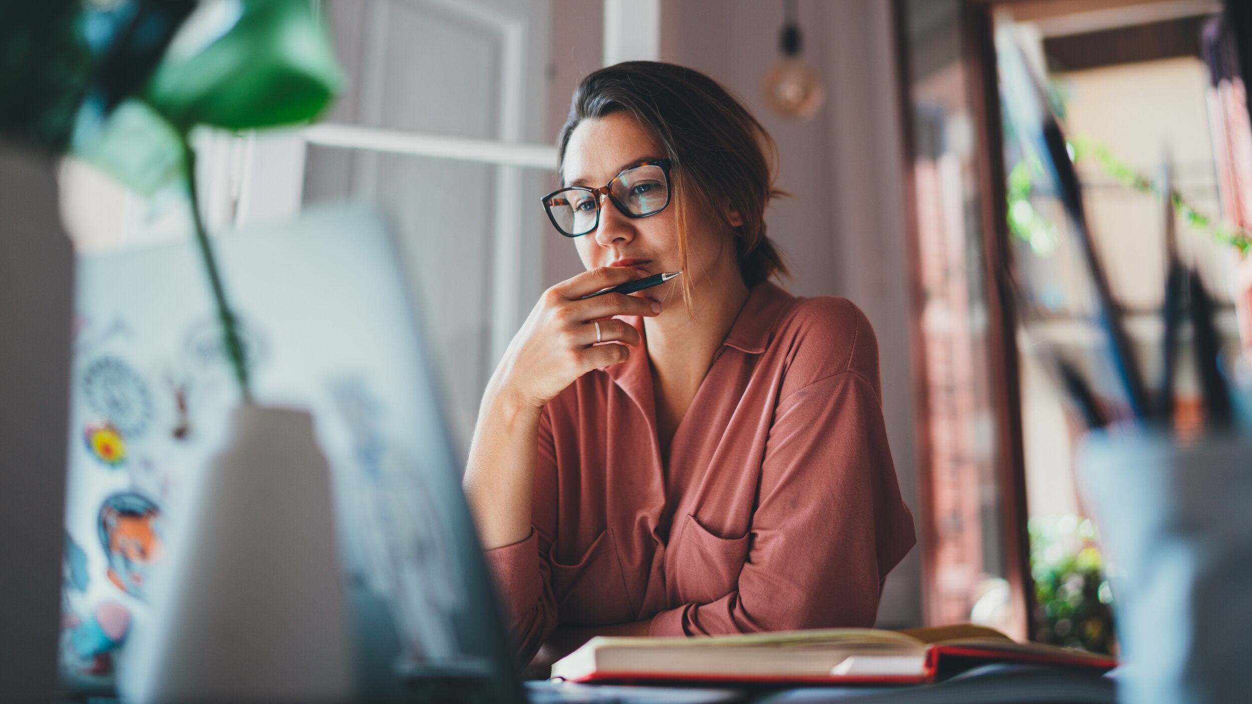 Woman thinking while looking at her laptop in a room at home. She wears a red shirt and glasses.