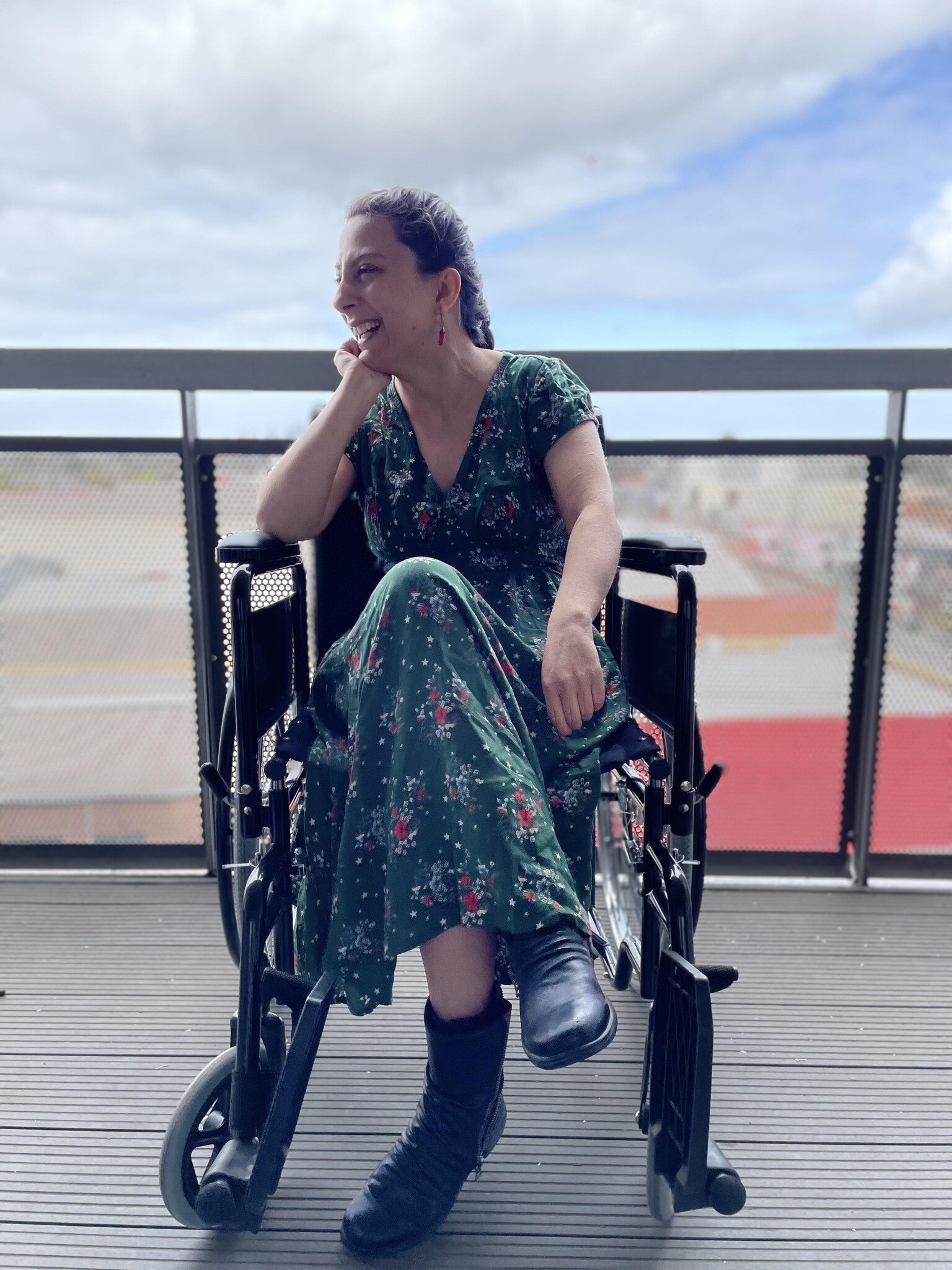 Laura wears a long green floral dress and black boots. She looks sideways and smiles, sitting in a wheelchair on a high balcony.
