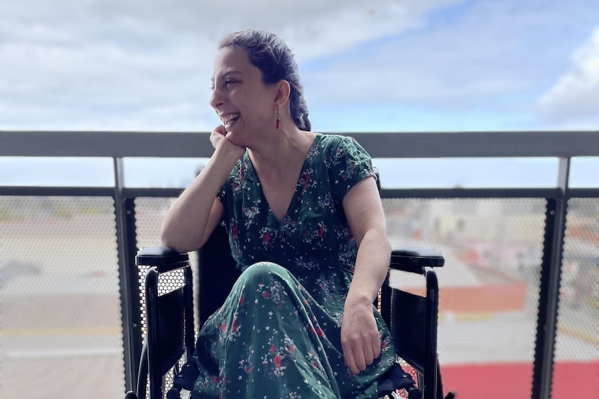 Laura wears a long green floral dress and black boots. She looks sideways and smiles, sitting in a wheelchair on a high balcony.