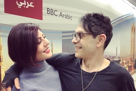 A man puts his arm around a woman and they smile at each other in front of a BBC Arabic sign