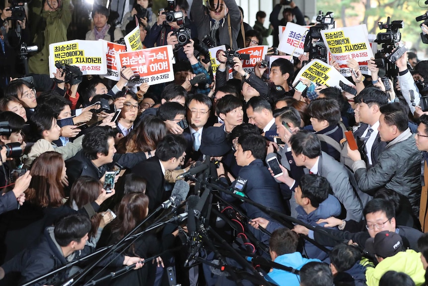 Choi Soon-sil surrounded by officers, media and protesters holding signs calling for her arrest in Seoul.