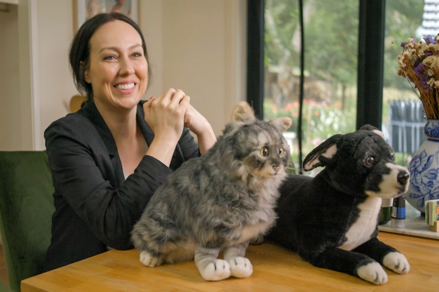A woman smiles with a stuffed cat and dog on a table in front of her.