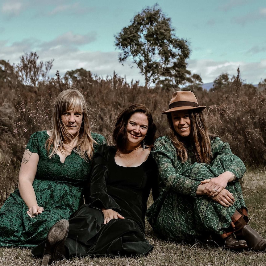 Three women wearing green dresses sit on the ground in the bush.