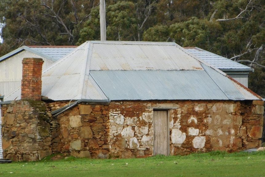 A stone building with a tin roof. It is bordered by green grass and trees are visible in the background.