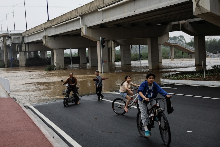 People ride bikes near a flooded road.