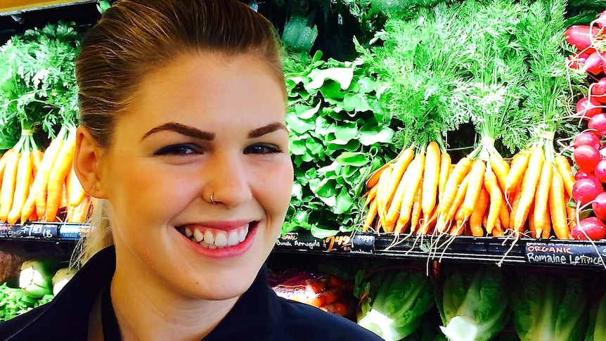 A smiling Belle Gibson stands next to fruit and vegetables in a shop.