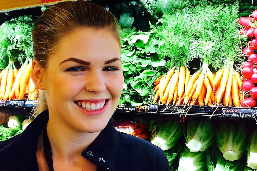 A smiling Belle Gibson stands next to fruit and vegetables in a shop.