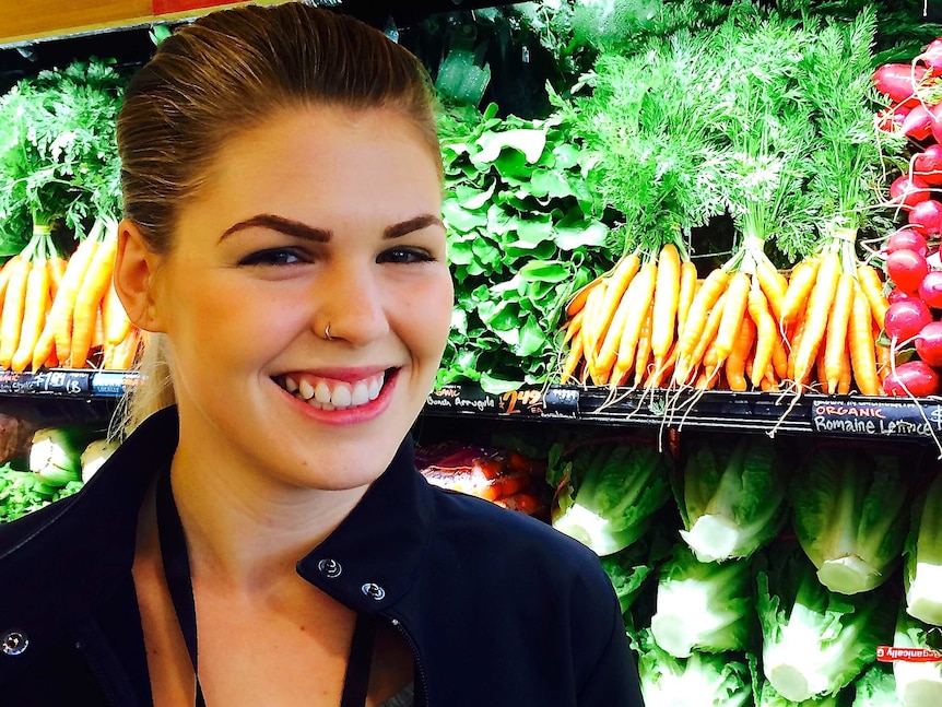 A smiling Belle Gibson stands next to fruit and vegetables in a shop.