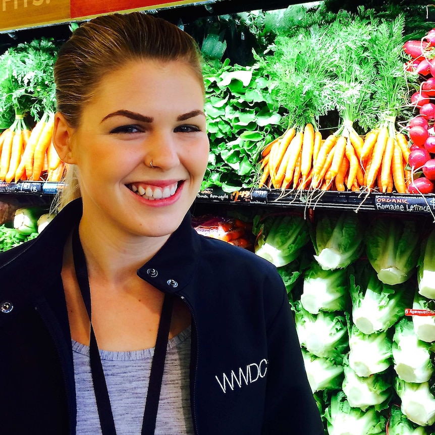 A smiling Belle Gibson stands next to fruit and vegetables in a shop.