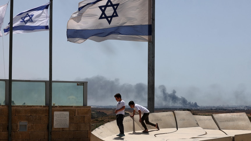 Two young boys in white T-shirts stand in the foreground underneath an Israeli flag as smoke rises from a town in the distance.