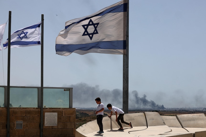 Two young boys in white T-shirts stand in the foreground underneath an Israeli flag as smoke rises from a town in the distance.