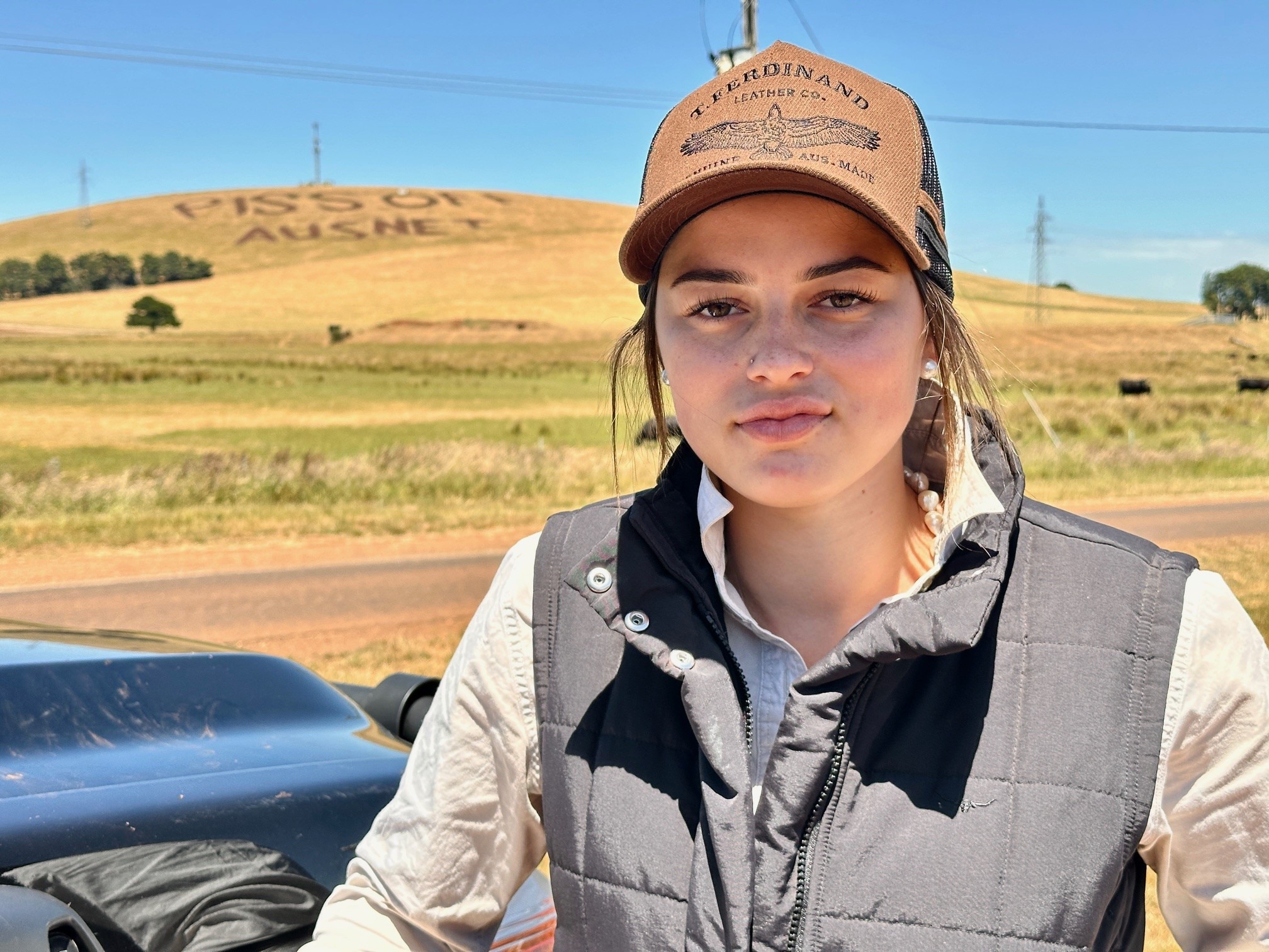A young woman wearing a cap, black vest and work shirt. In the distance is a green hill that reads 'Piss off Ausnet'.