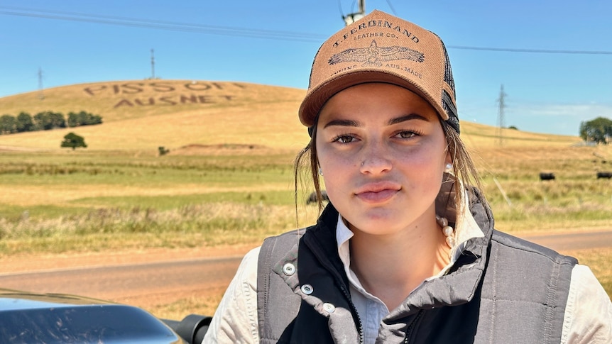 A young woman wearing a cap, black vest and work shirt. In the distance is a green hill that reads 'Piss off Ausnet'.