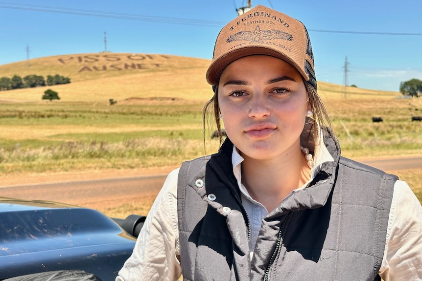A young woman wearing a cap, black vest and work shirt. In the distance is a green hill that reads 'Piss off Ausnet'.
