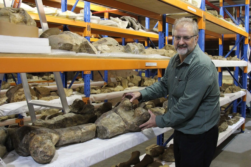 Man wearing spectacles stands beside shelves of dinosaur bones