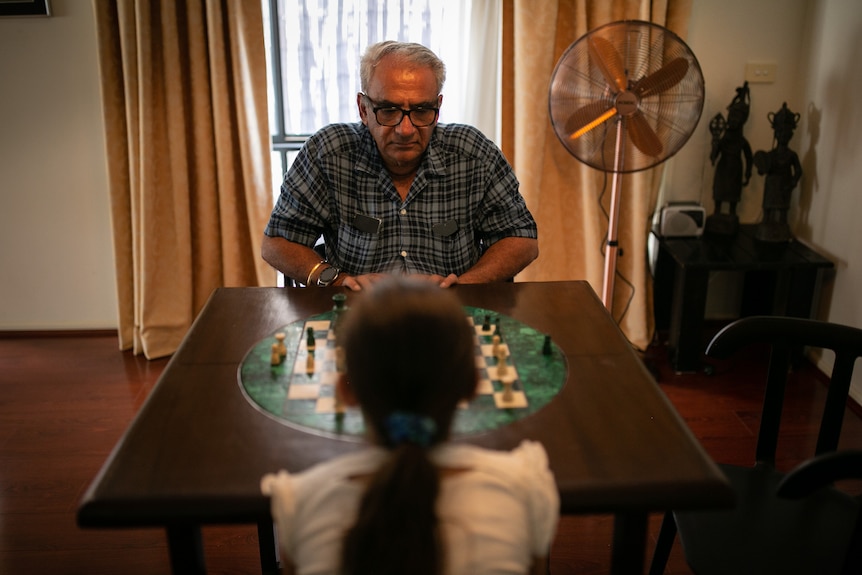 A man with grey hair sits across a chess board from a young girl, with a fan in the background.