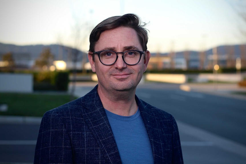 A clean-shaven man in spectacles and a casual jacket stands outside, smiling at the camera.