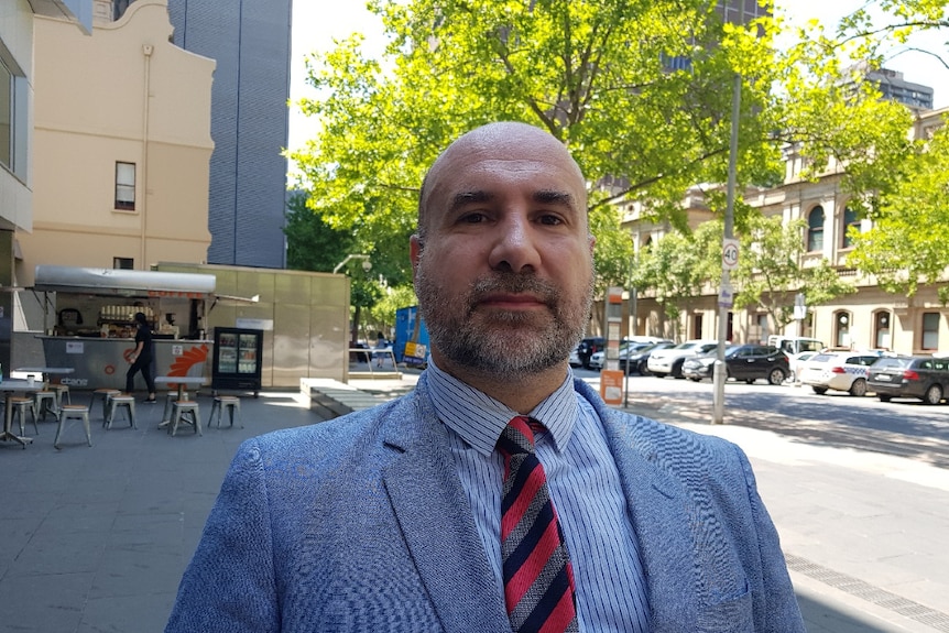 Mark Stanarevic wears a suit and tie with a neutral expression on his face as he stands outside in an urban area.