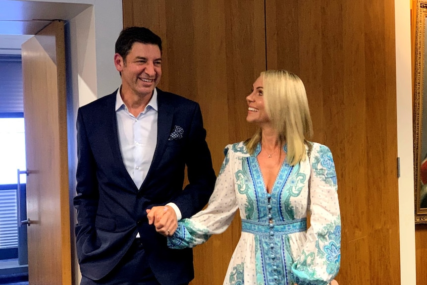 Basil Zempilas wearing a suit and his wife Amy wearing a white and blue dress hold hands and smile.