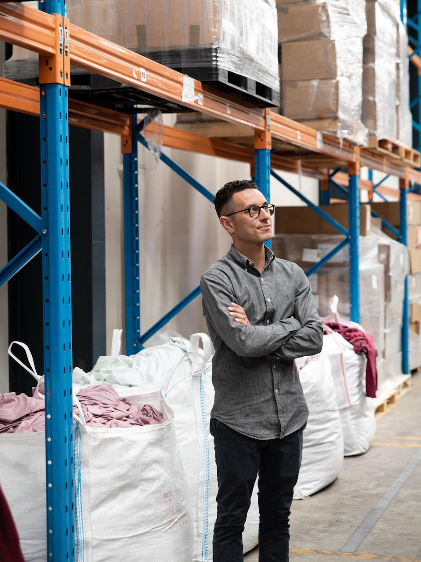 A man in a business shirt stands in front of warehouse shelves with bags of clothes