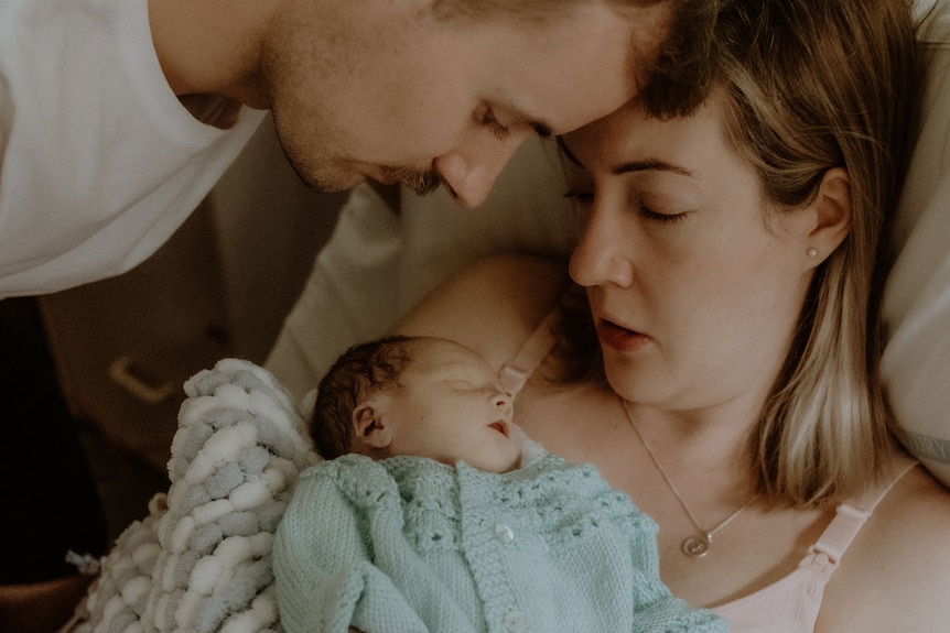 Dimity in a hospital bed, holds baby Finley while Michael looks over them.