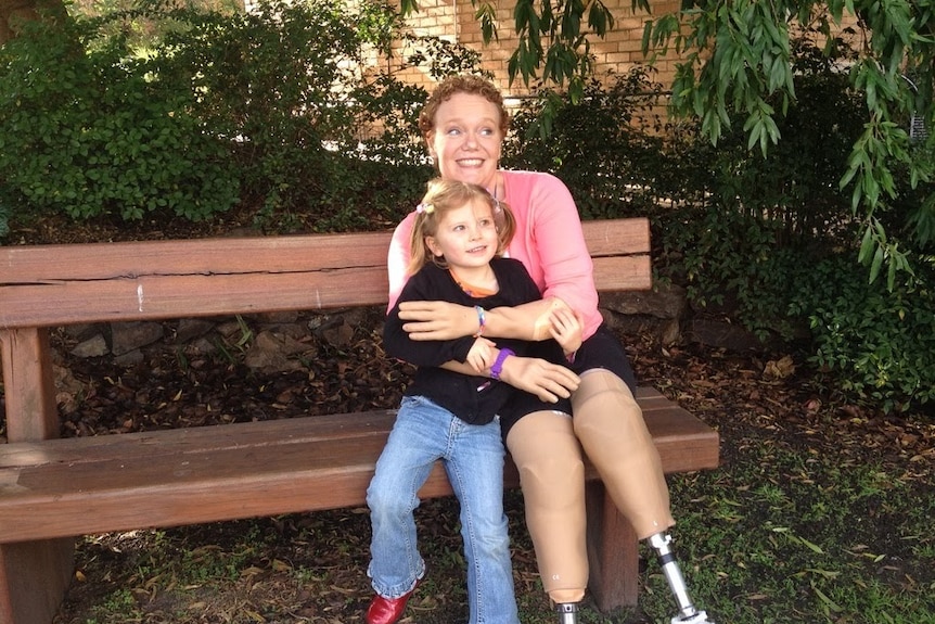 A woman with short, curly hair and prosthetic arms and legs sits on a parl bench smiling and hugging her daughter.