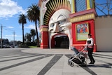 A woman wearing a face mask and pushing a pram walks past Luna Park in St Kilda on a sunny day.