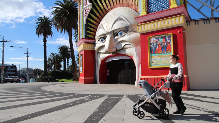 A woman wearing a face mask and pushing a pram walks past Luna Park in St Kilda on a sunny day.