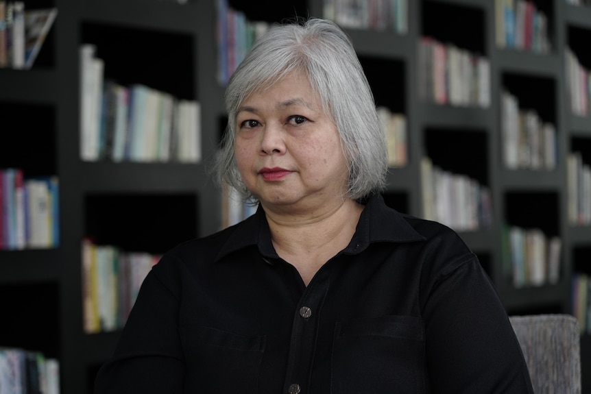 A middle-aged woman stands in front of a bookcase, wearing a black buttoned shirt.